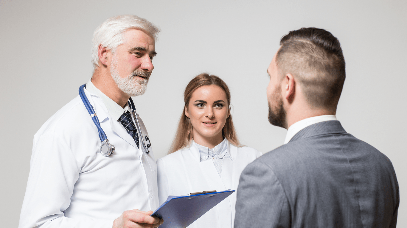 a imagem mostra um médico de cabelos brancos, muito atencioso, conversando com o paciente, que é um homem de terno e cabelos negros. O médico está falando sobre a importância dos 3 pilares estratégicos da Experiência do Paciente.